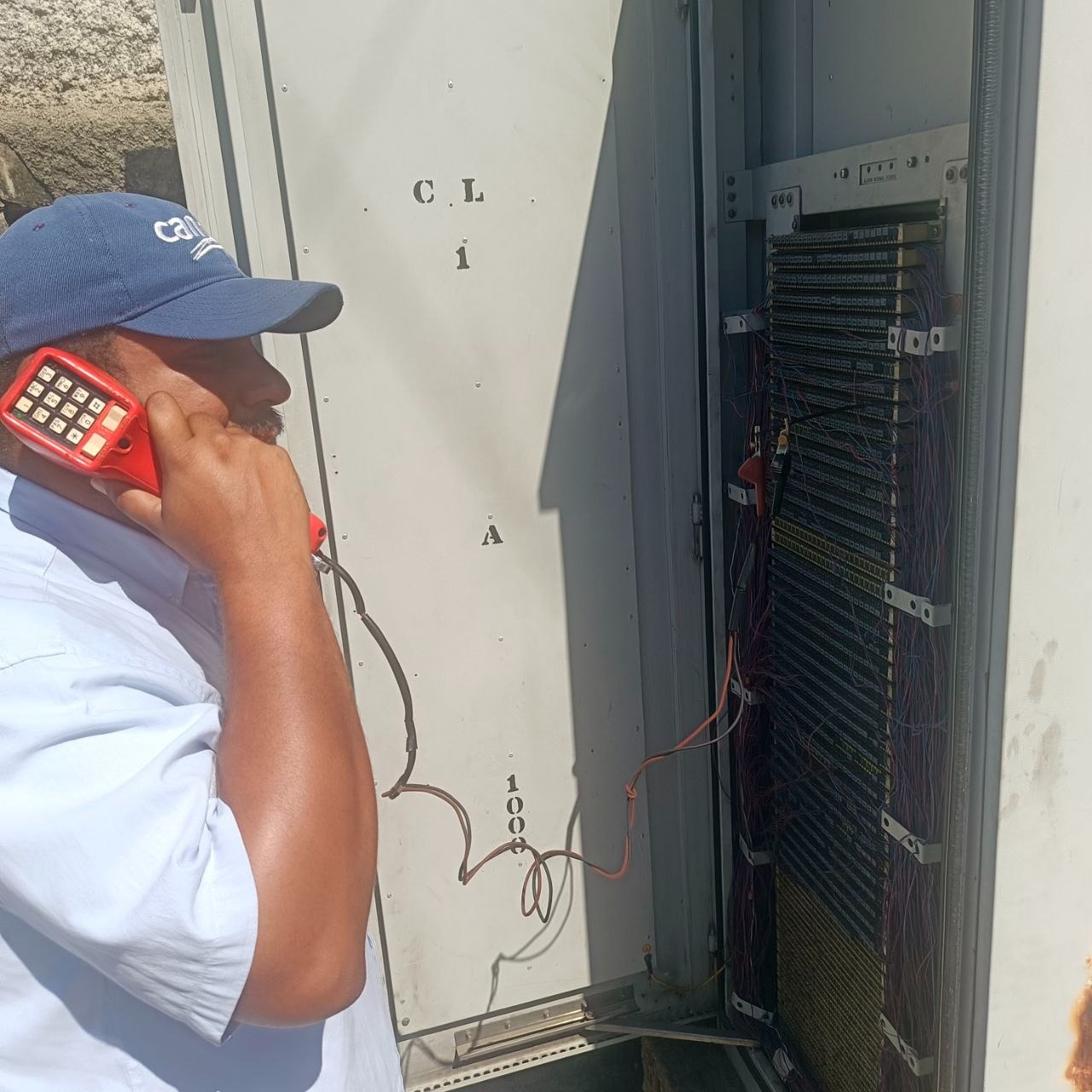 Técnico de Cantv con un teléfono de prueba rojo al oído frente a un armario de conexiones telefónicas abierto