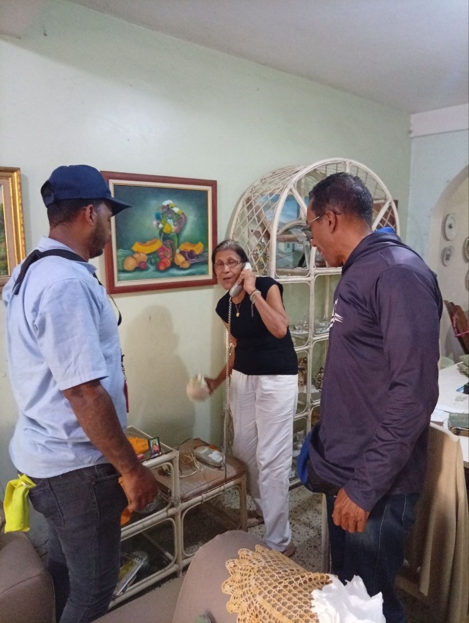 Dos hombres, uno con gorra azul y camisa azul claro, en la sala de una casa mientras una mujer habla por un teléfono fijo de cable