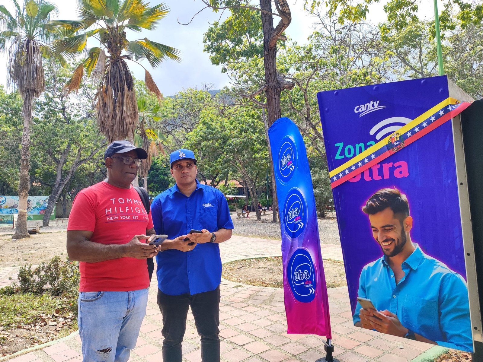 Pendón de Zona Ultra de Cantv en una plaza con palmeras, junto a dos hombres que revisan un teléfono